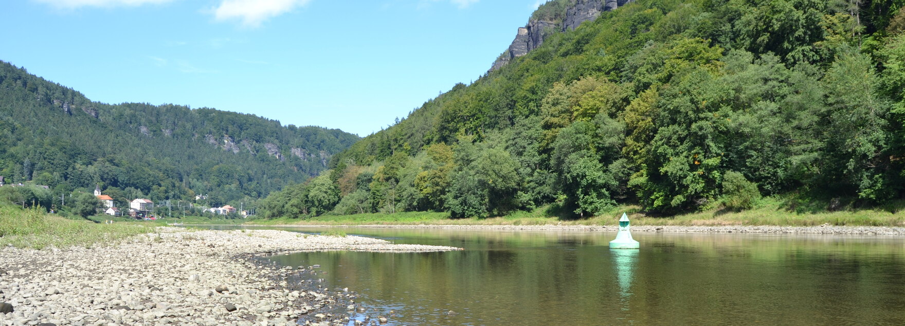 Ein Fluss, welcher nach vorne hin breiter wird. Am linken Ufer befinden sich trocken gefallene Kieselbänke. Manche davon ragen bis in die Mitte des Flusses hinein.